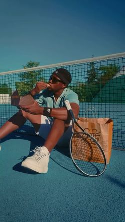 Un jeune homme assis sur un court de tennis bleu, mangeant un sandwich avec une raquette et un sac en papier à côté.
