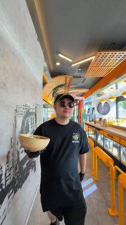Serveur portant un chapeau et lunettes, tenant un bol, dans un restaurant asiatique moderne avec décor jaune.