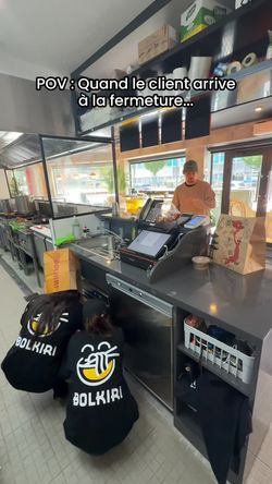 Deux employés en pulls BOLKIRI travaillent derrière le comptoir d'un restaurant asiatique avec caisse et sacs à emporter.