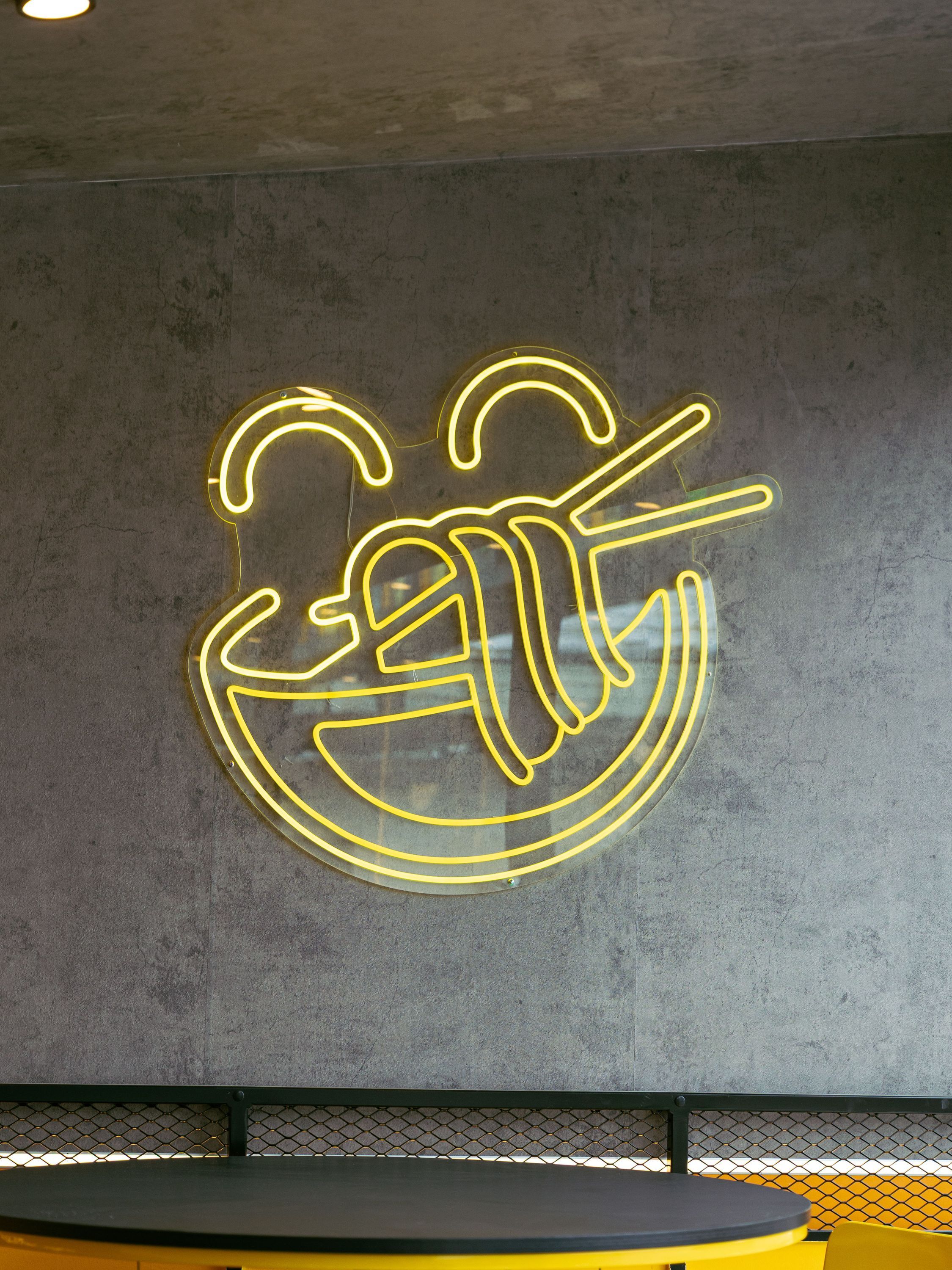 Néon jaune représentant un bol de nouilles avec des baguettes sur un fond mural gris dans un restaurant asiatique.