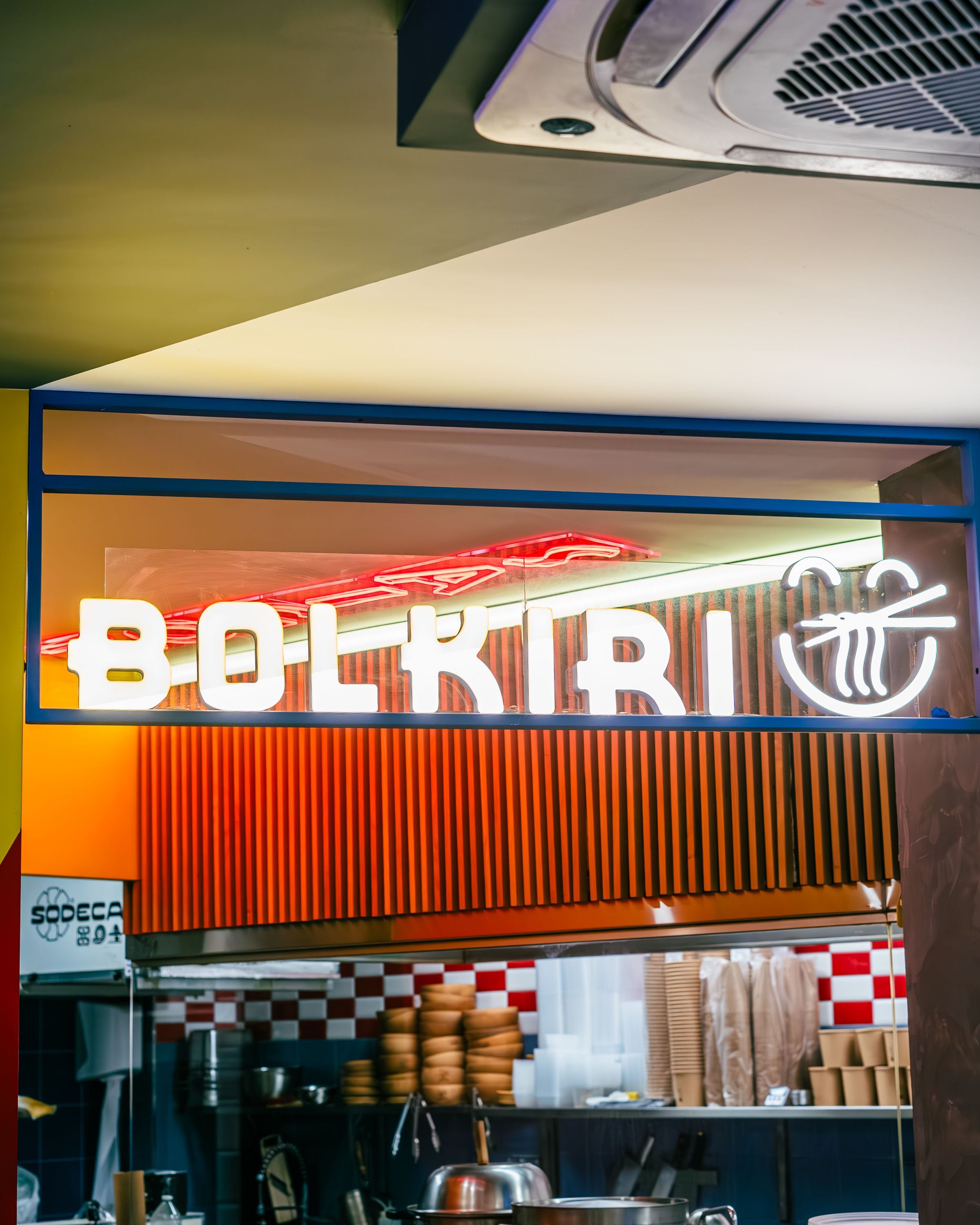 Enseigne lumineuse BOLKIRI éclairant l'entrée du restaurant asiatique avec cuisine visible en arrière-plan.