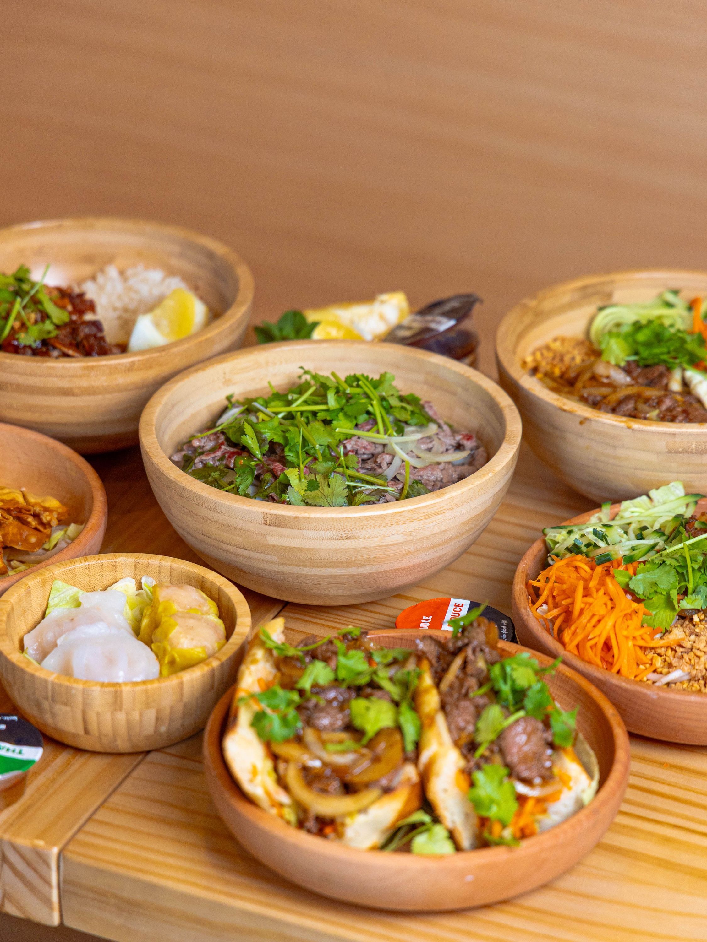 Plats variés asiatiques servis dans des bols en bois avec herbes fraîches et légumes colorés.