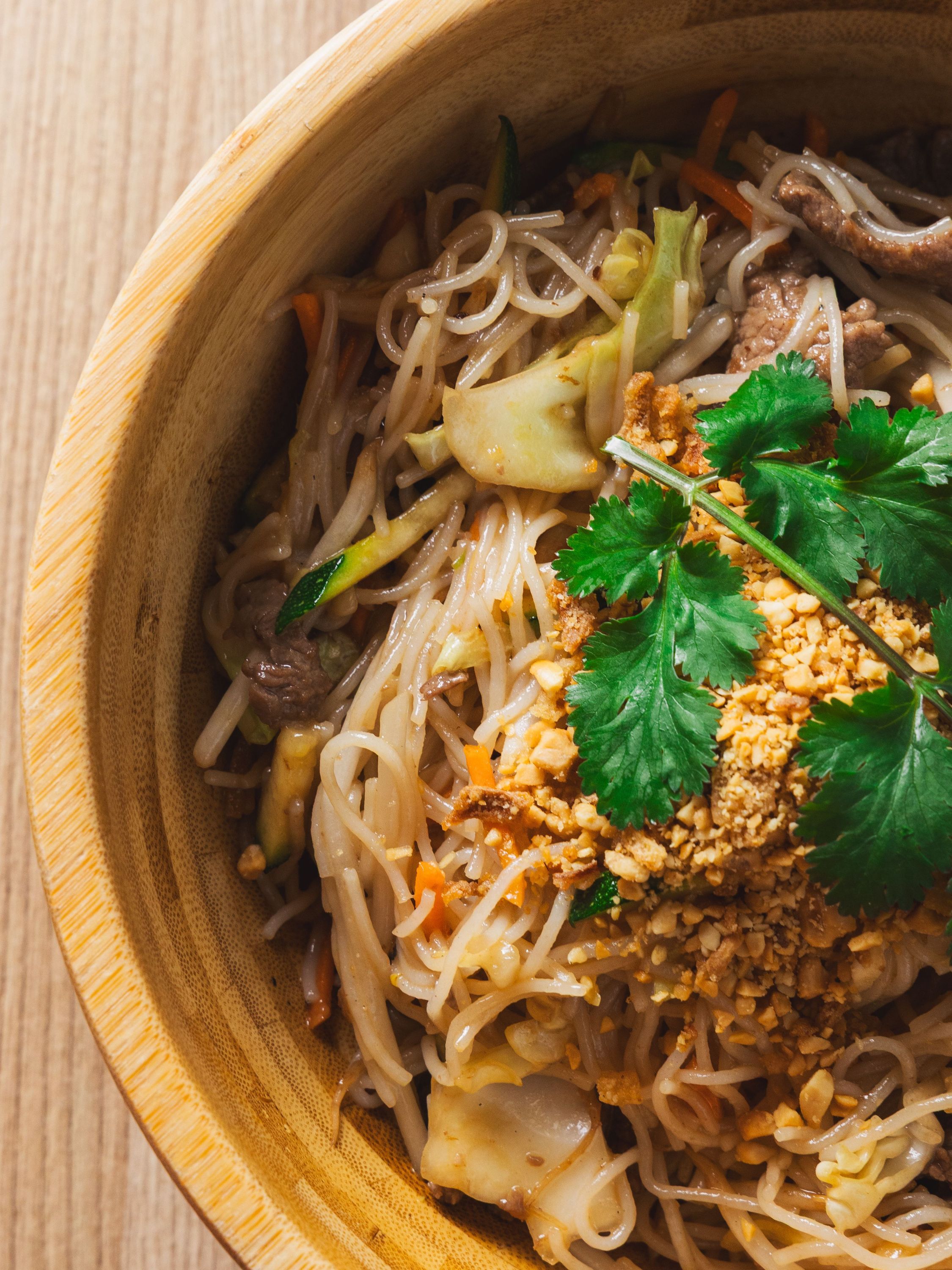 Plat asiatique dans un bol en bois avec nouilles, légumes, viande, coriandre et cacahuètes concassées
