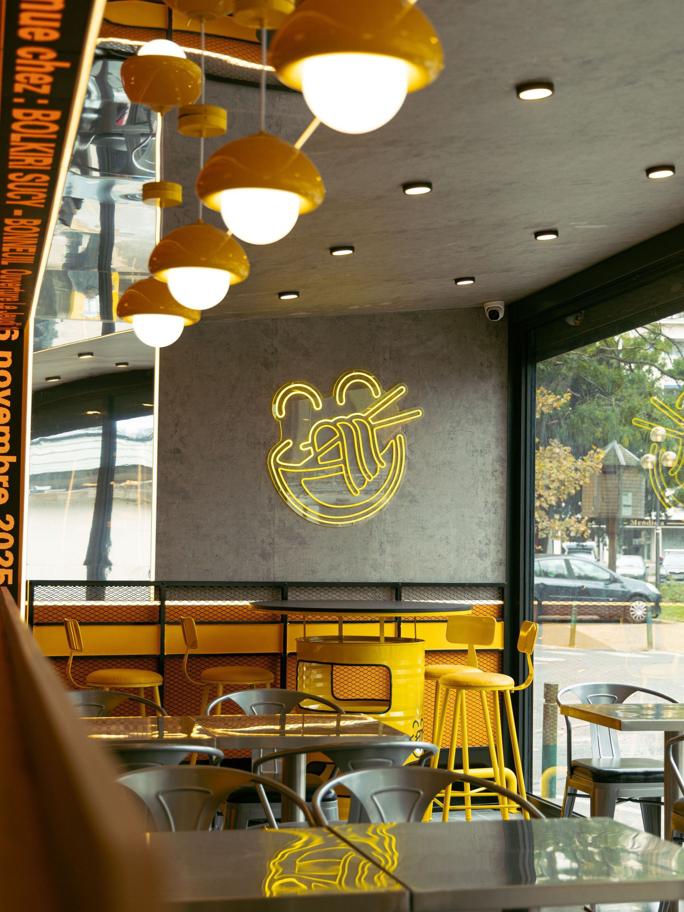Restaurant asiatique avec éclairage moderne jaune, tabourets hauts et mur décoré d'un néon en forme de bol de nouilles.