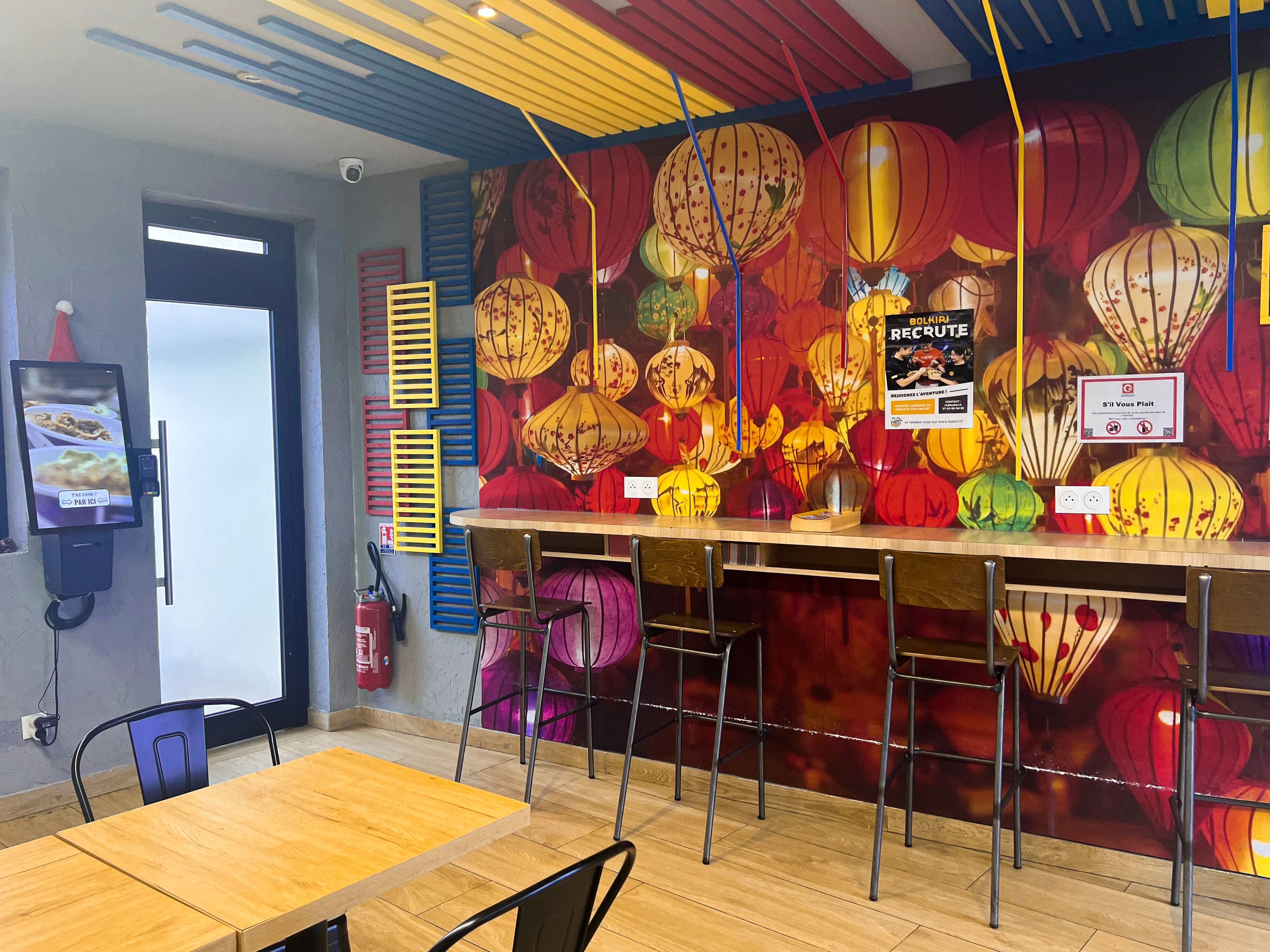 Espace intérieur coloré avec des lanternes, tables en bois et chaises, ambiance street food asiatique.