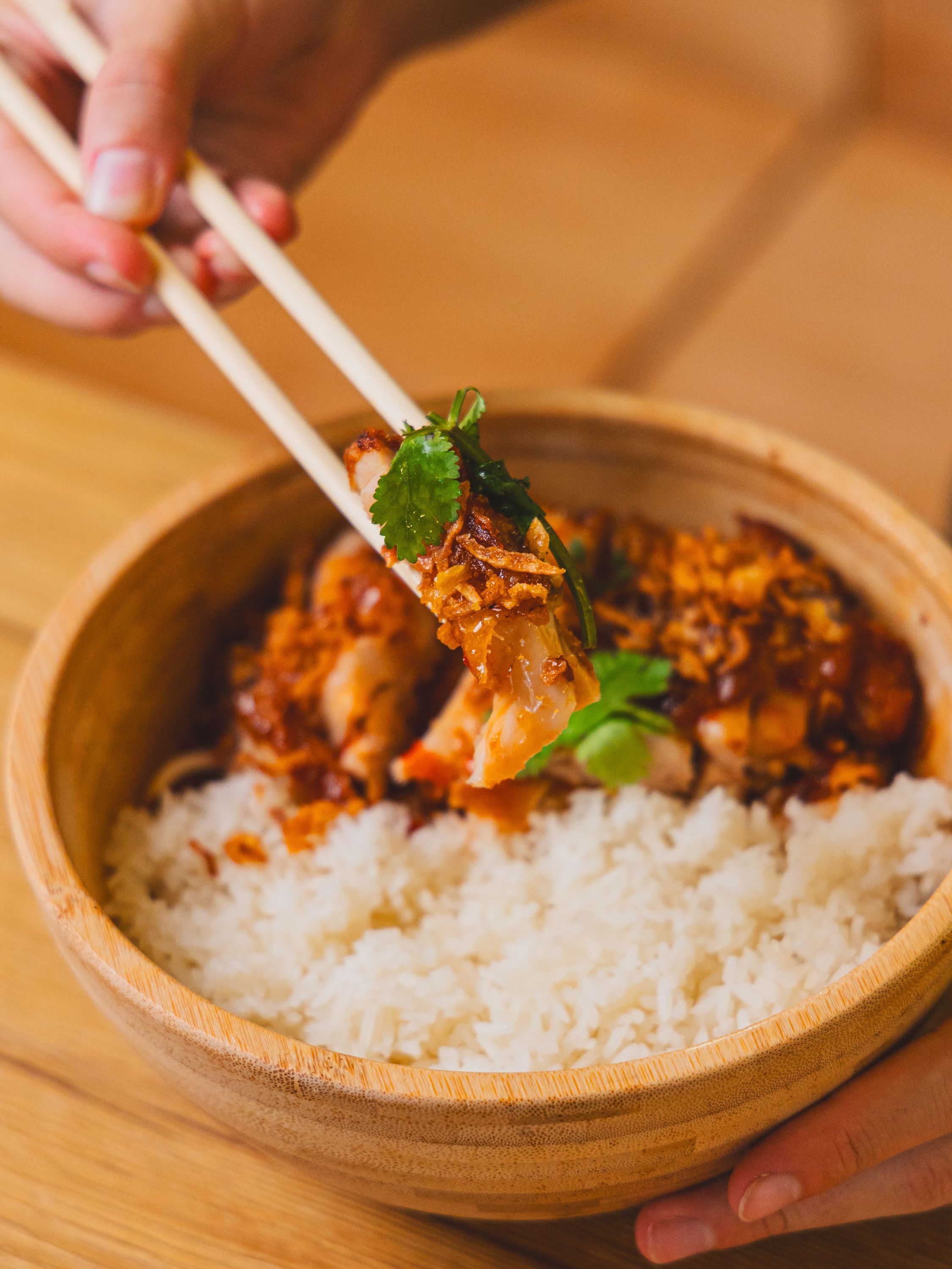 Bol en bambou avec riz blanc et poulet asiatique garni de coriandre et oignons frits, baguettes tenues à la main.
