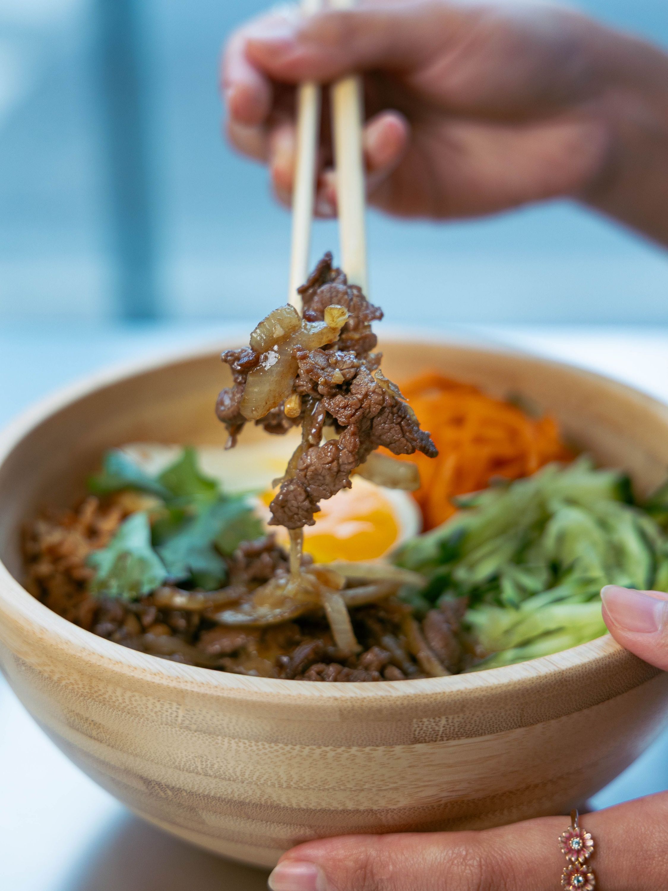 Plat asiatique dans un bol en bambou avec viande, légumes, carottes râpées et œuf, tenu par des baguettes.
