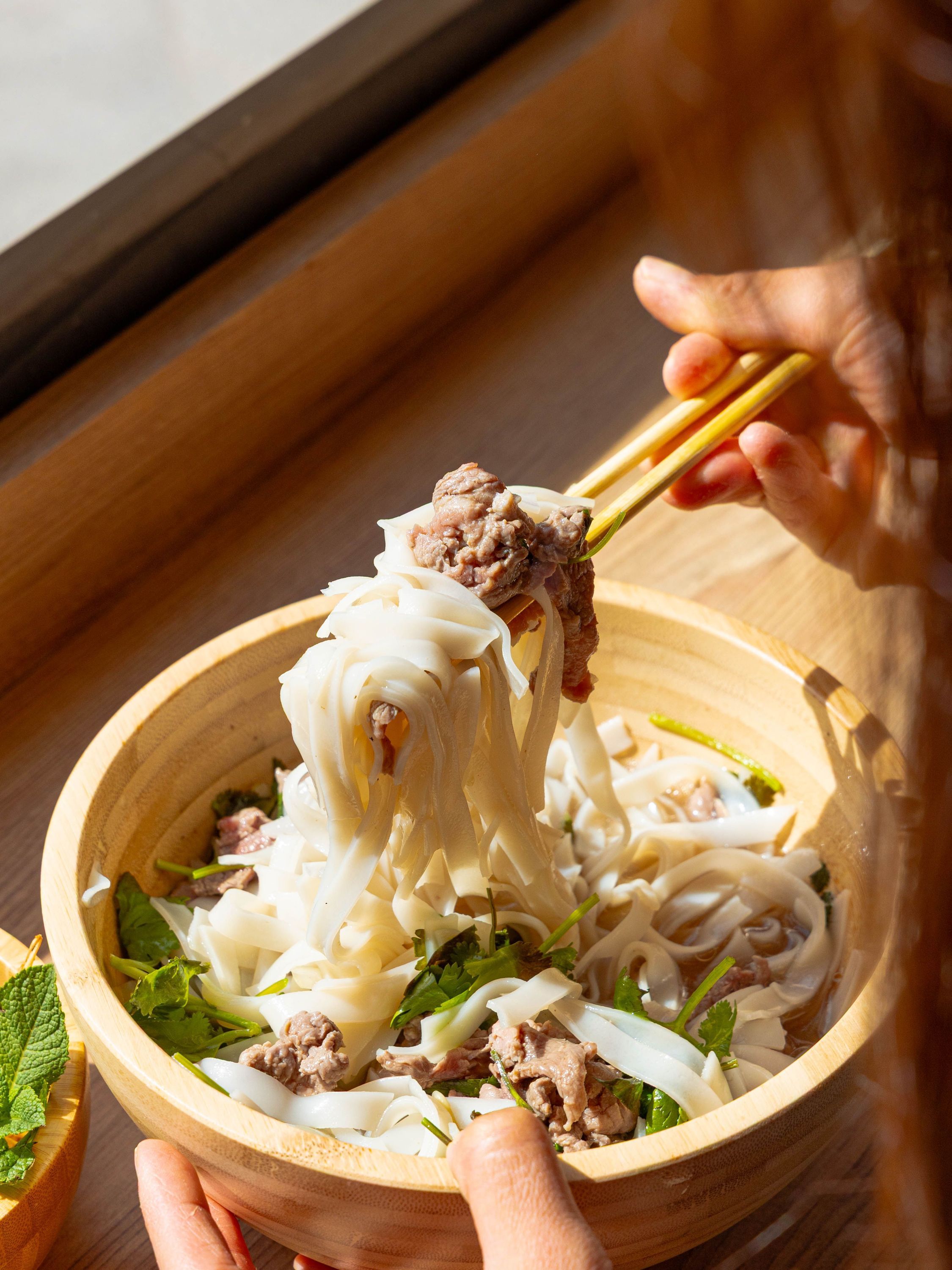 Plat asiatique de nouilles plates avec viande et herbes fraîches servi dans un bol en bois.