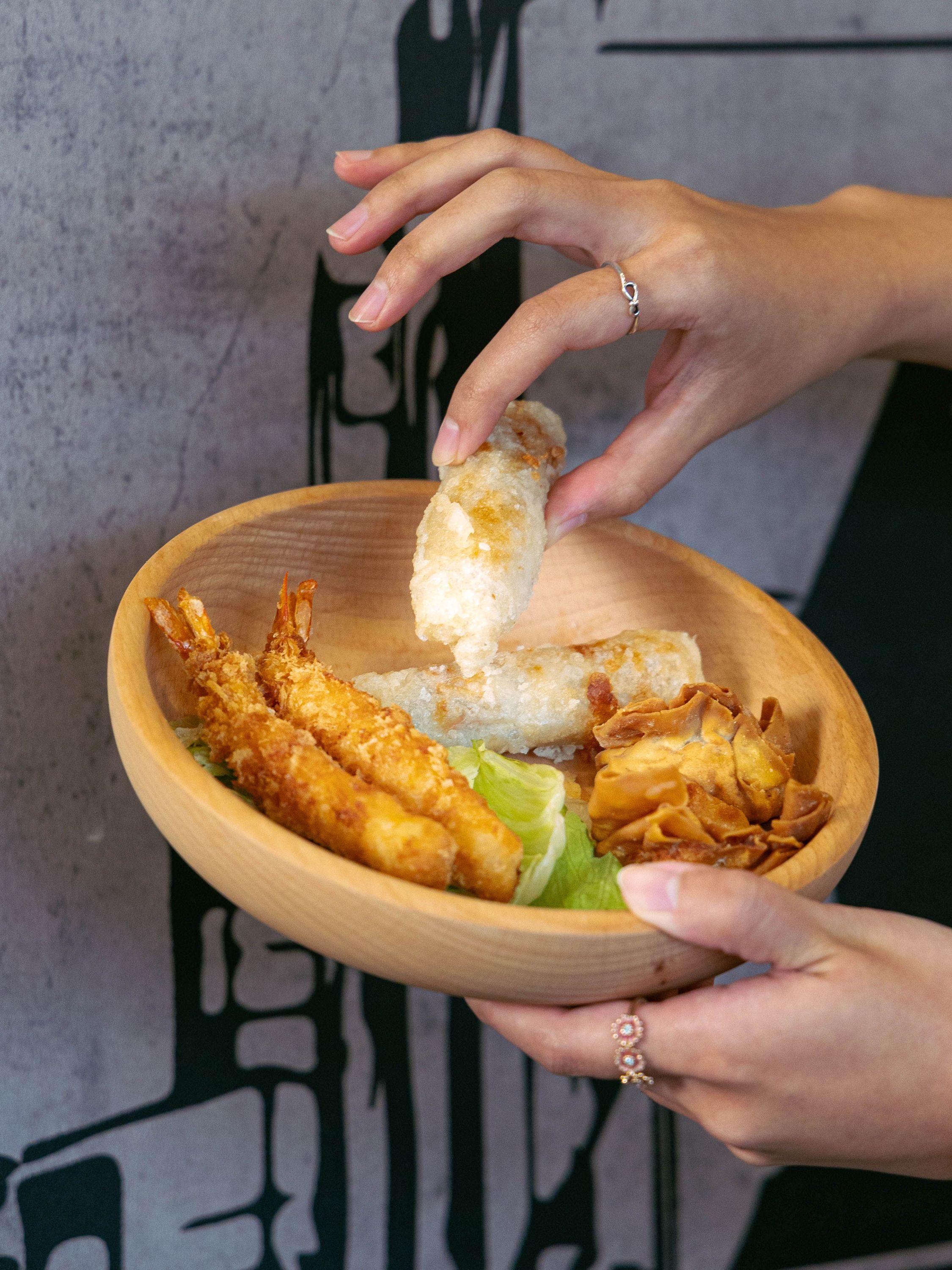 Main tenant un bol en bois contenant crevettes frites, rouleaux de printemps et chips dans un restaurant asiatique.