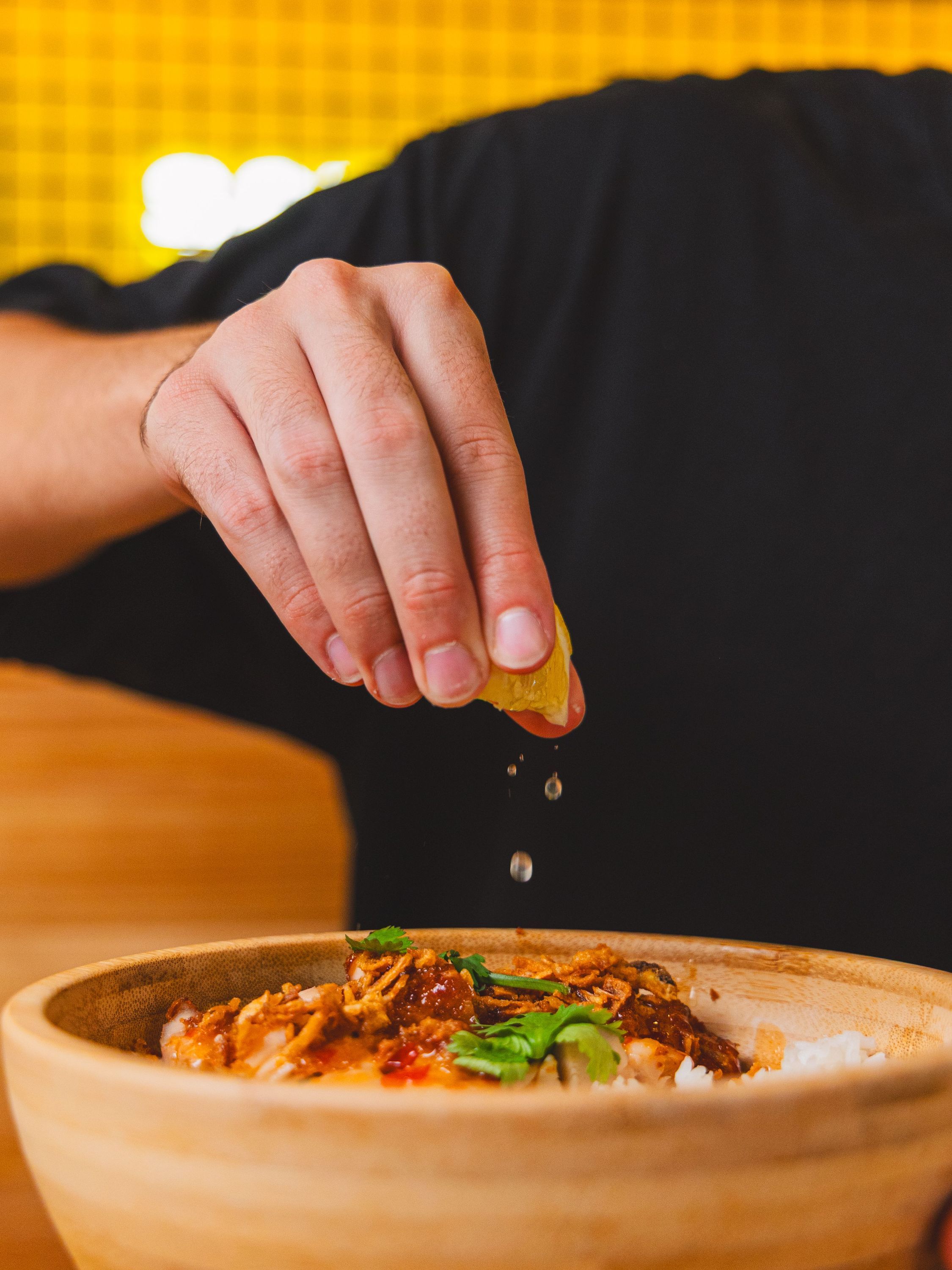 Un plat asiatique dans un bol en bois avec un citron pressé au-dessus, gouttes de jus visibles, main en action